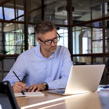 Man sitting at a desk looking at a laptop and taking notes