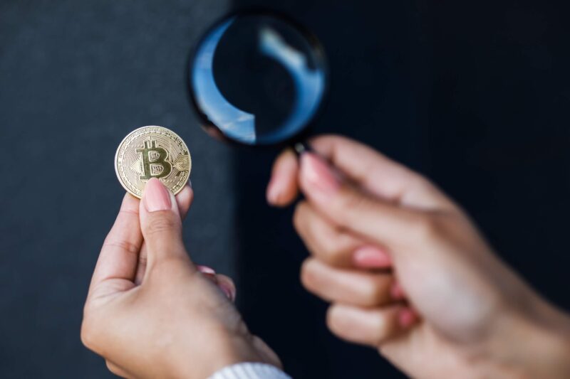 Hands holding Bitcoin and a magnifying glass